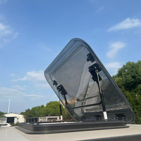 A transparent roof vent with black metal hinges and support brackets, shown open on a roof against a blue sky with clouds and green trees in the background.